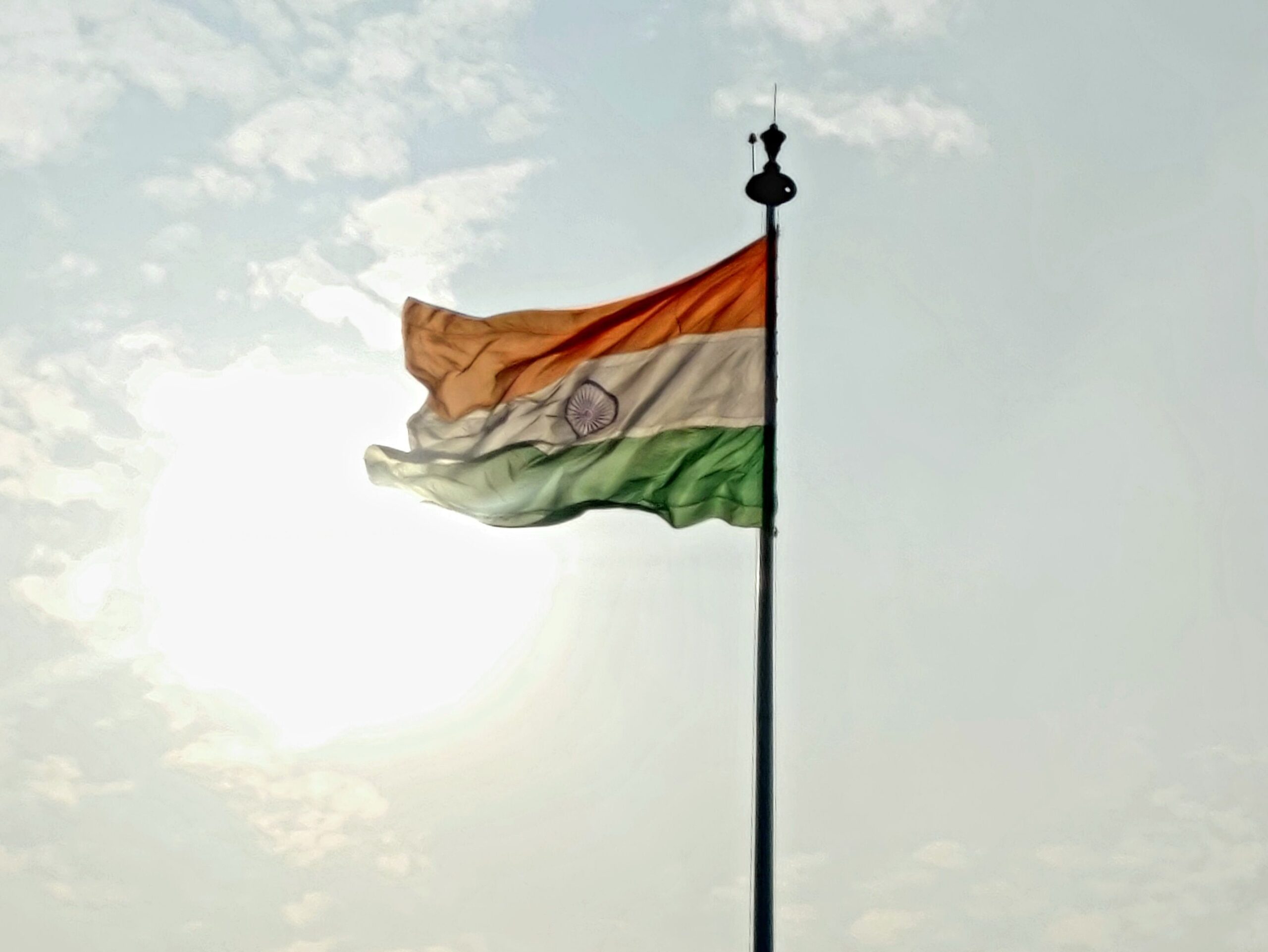 Indian national flag waving on flagpole against a bright sky with clouds.